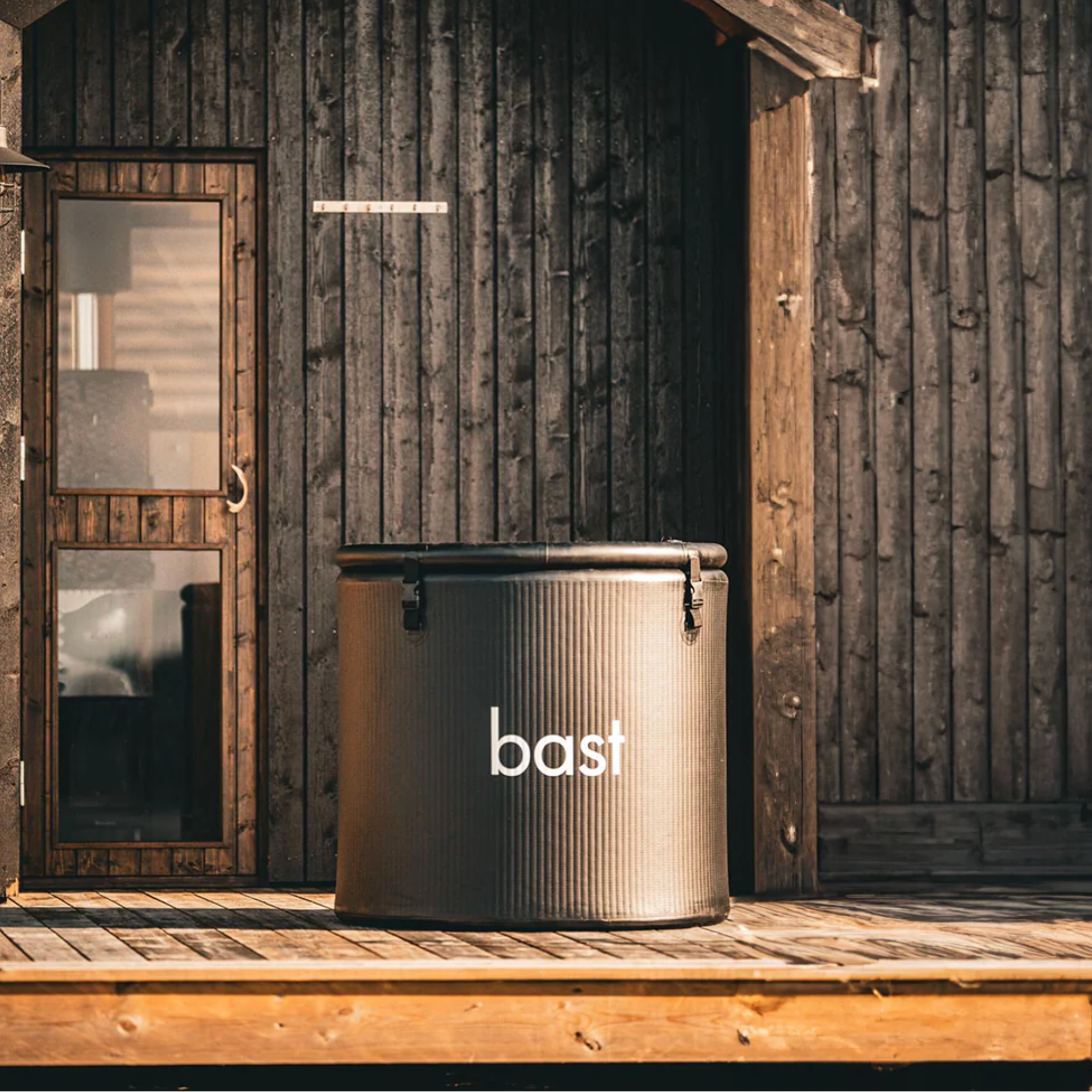 Black cylindrical inflatable ice bath with 'bast' branding in front of a wooden building