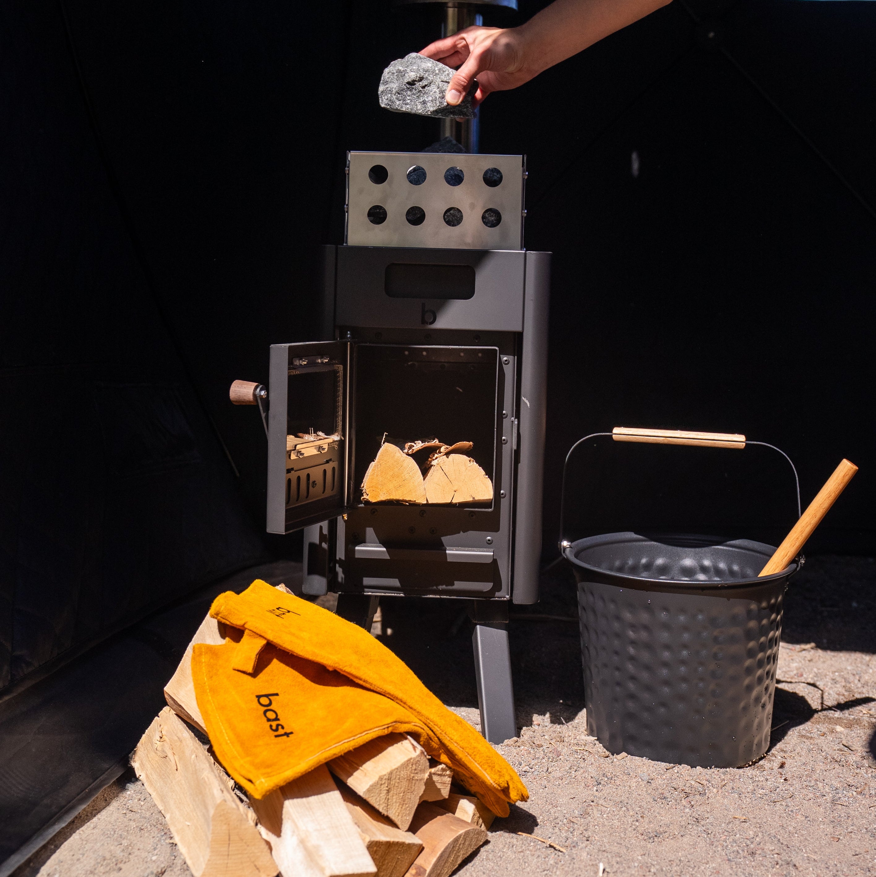 Person using a wood stove with firewood and a black bucket nearby