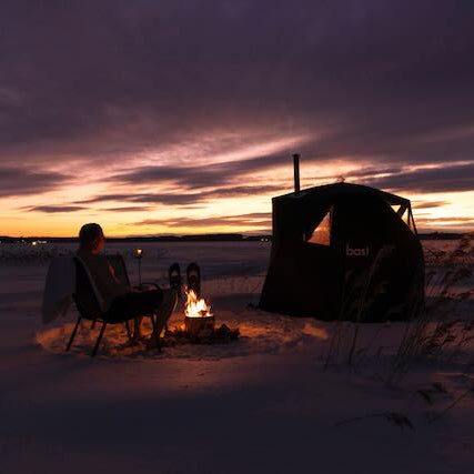 Person sitting by a campfire in a snowy landscape at sunset.