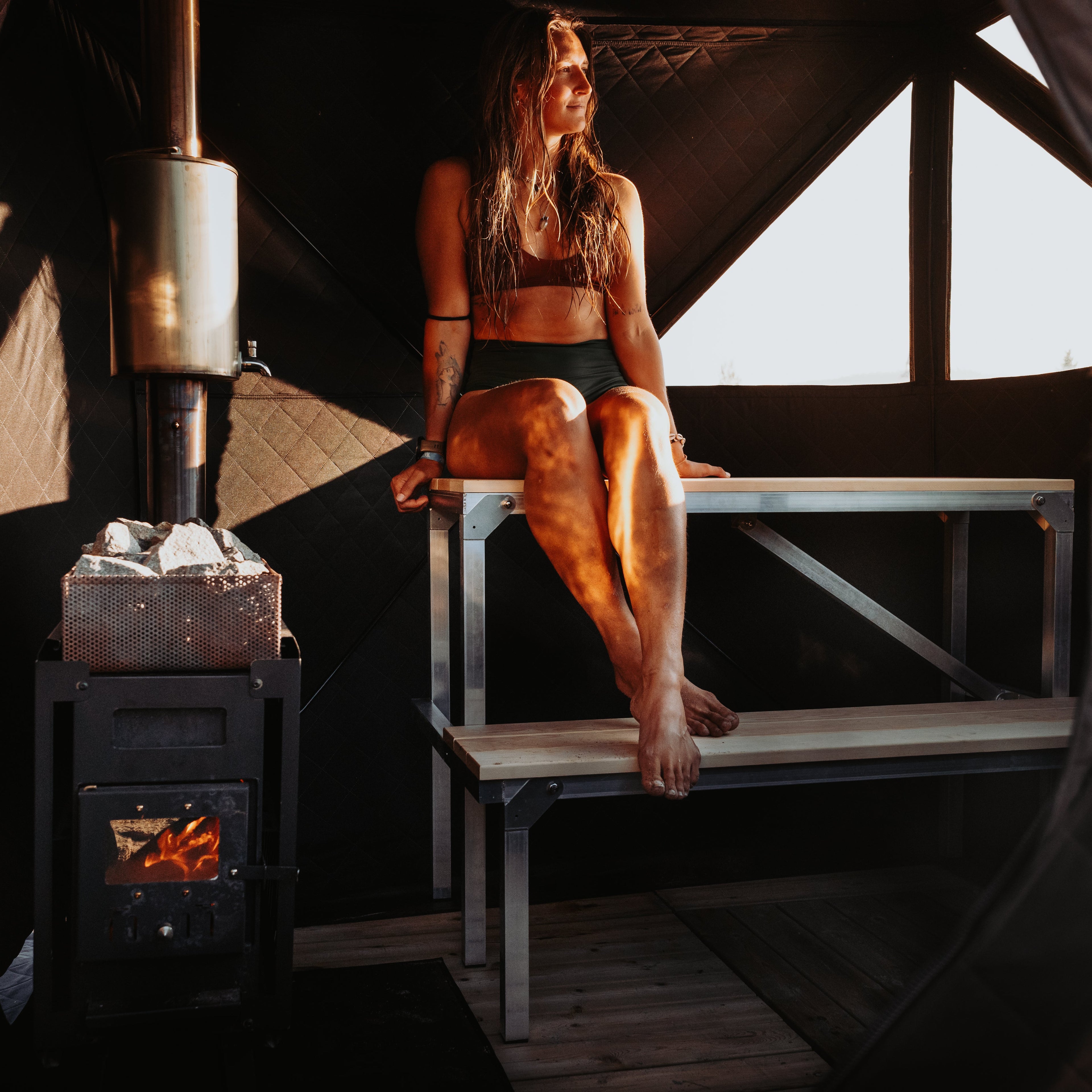 Person sitting on a bench in a sauna tent chamber with a wood stove.
