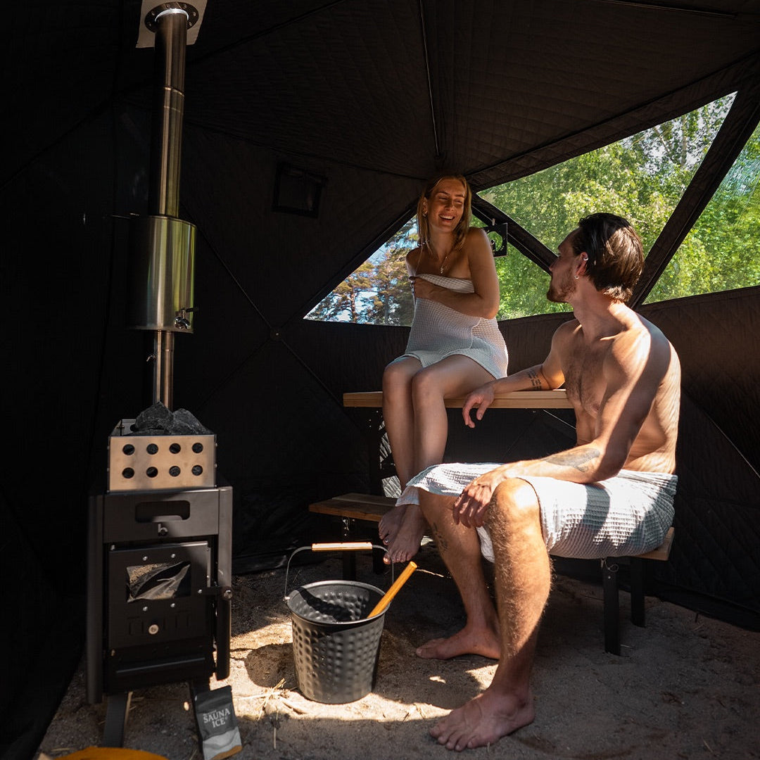 Two people sitting inside a sauna tent with a wood stove and equipment.