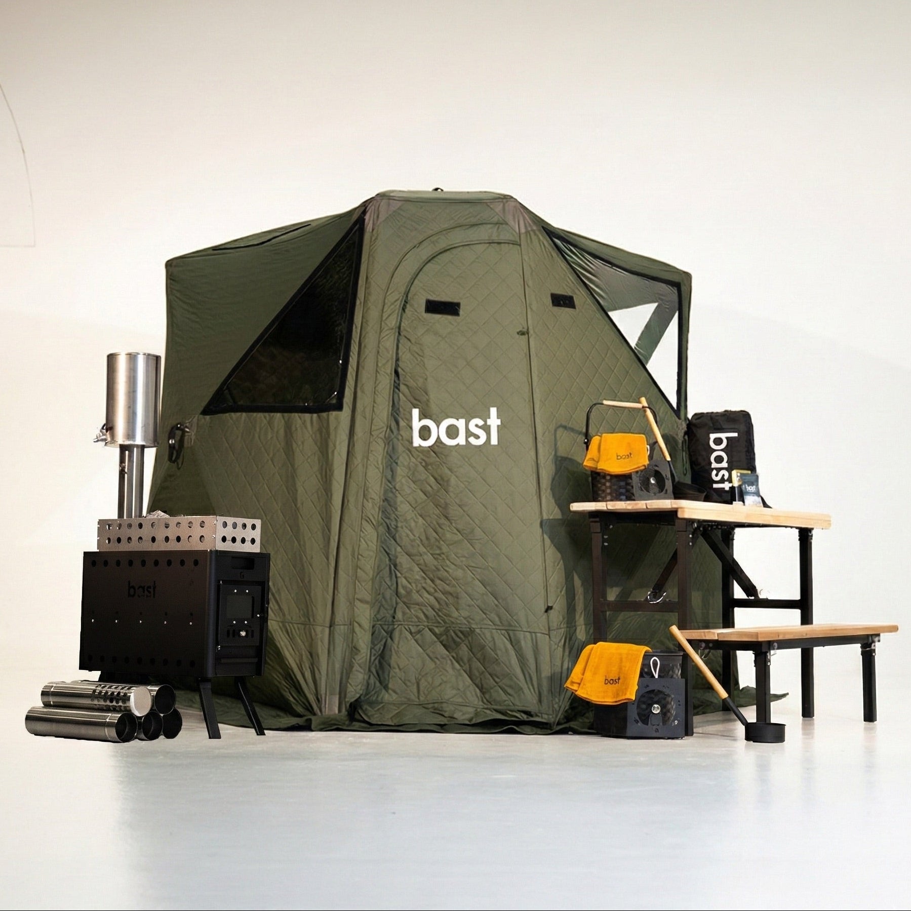 Green sauna tent with 'bast' branding, surrounded by sauna stove equipment and  a wooden bench on a white background