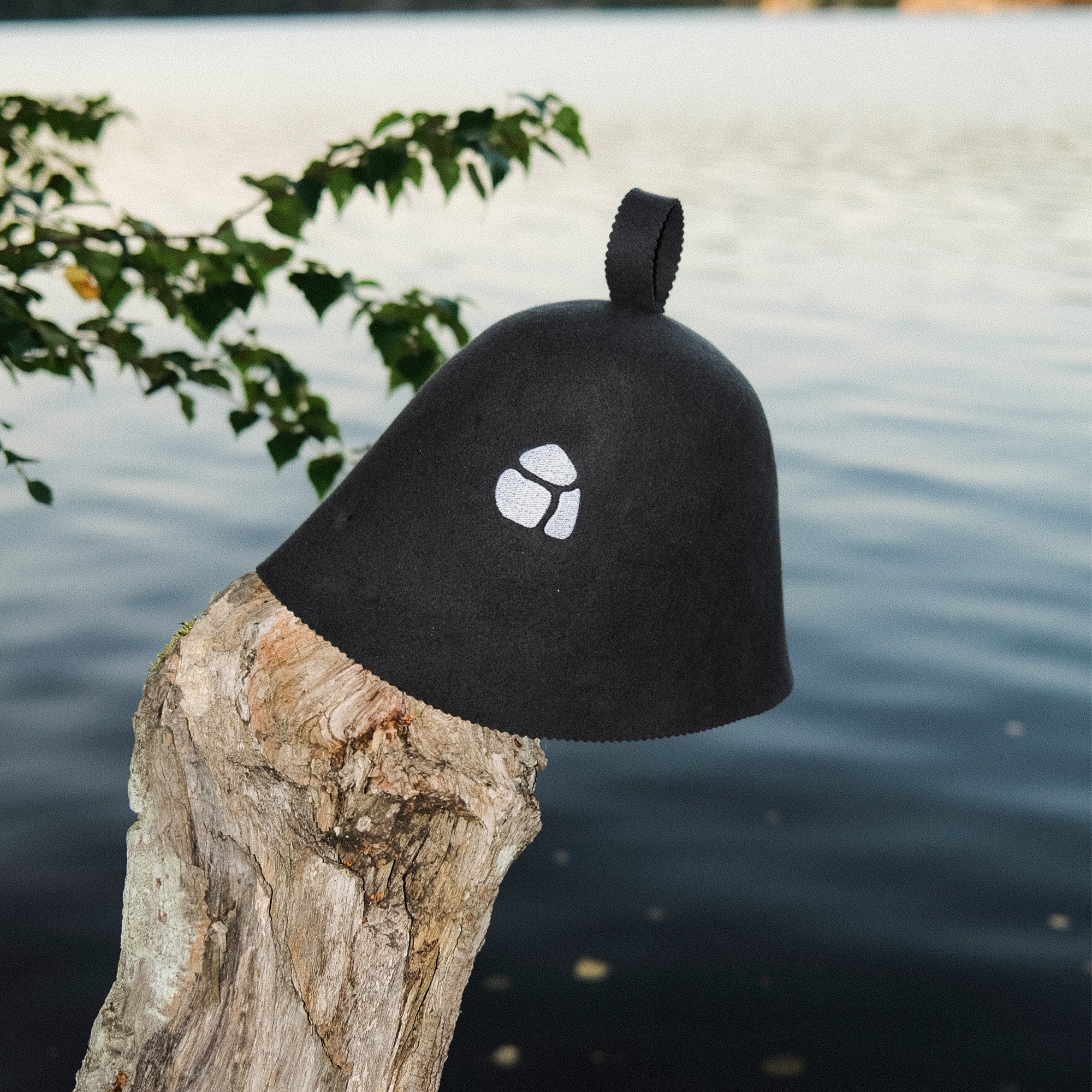grey sauna hat with a white embroidered logo on a wooden log by a body of water