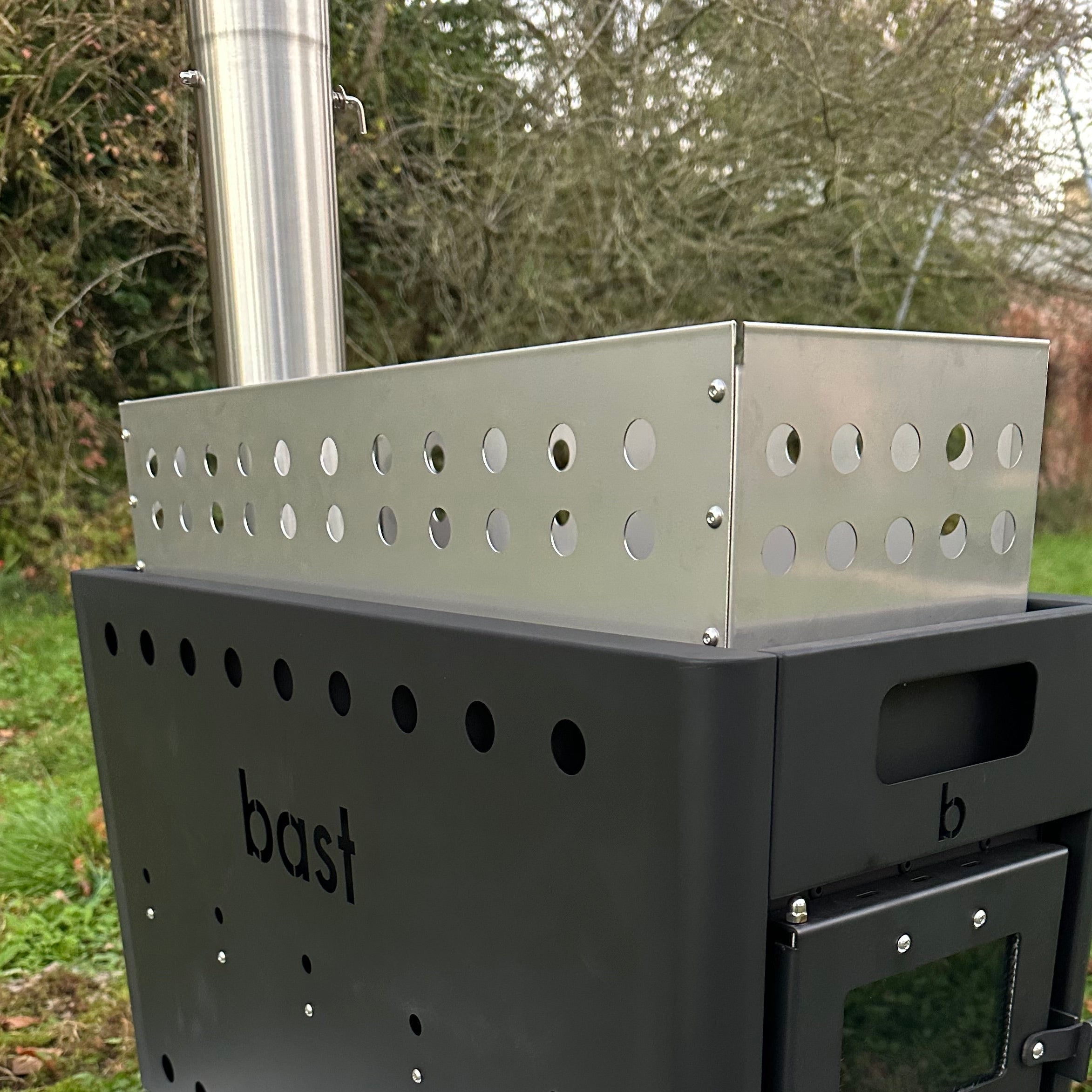sauna stone metal basket sits on top of a black stove with 'bast' branding in a outdoor enviroment