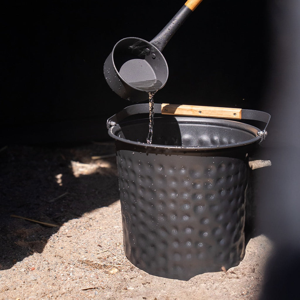 Person pouring water into a black bucket on a sandy surface.