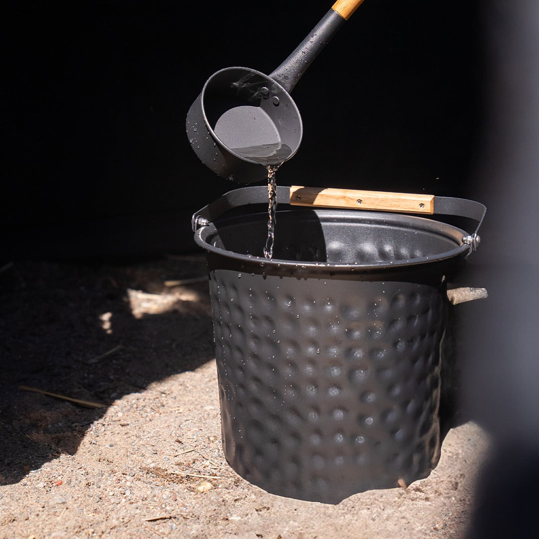 Person pouring water into a black bucket on a sandy surface.