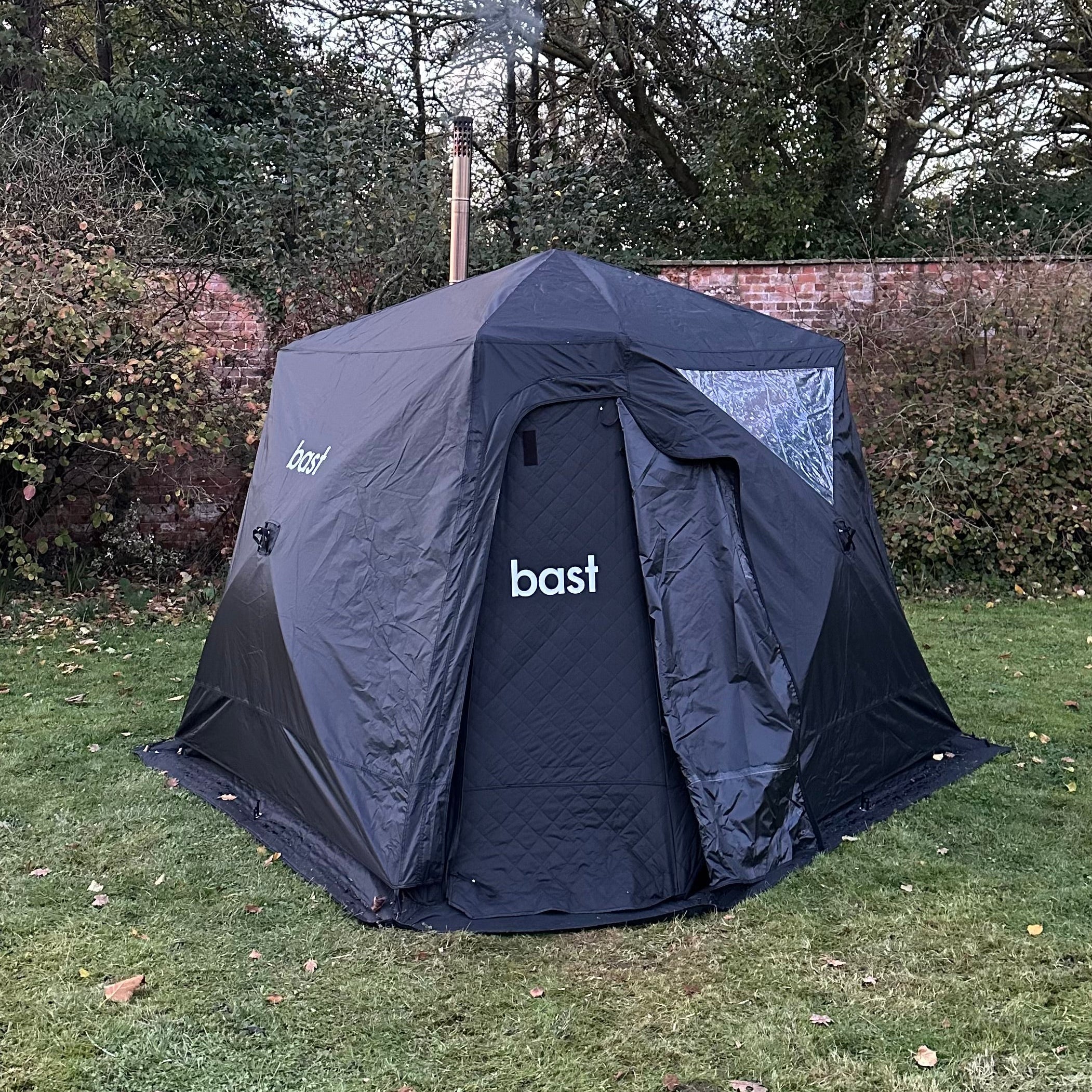 Black sauna tent with 'bast' branding on a grassy area with trees in the background